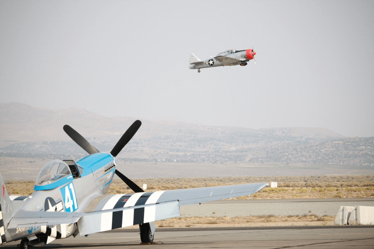 A P-51 Mustang is parked on the ground while a T-6 Texan flies past overhead