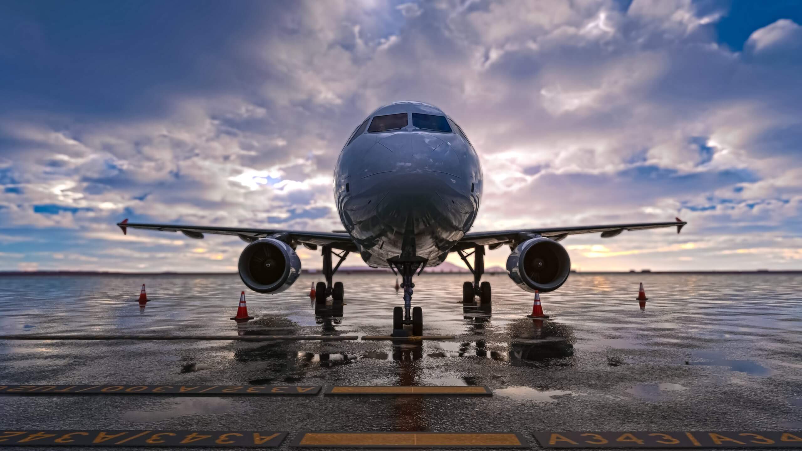 A front view of a large airplane on a wet tarmac. The sun rises in the distance behind it.