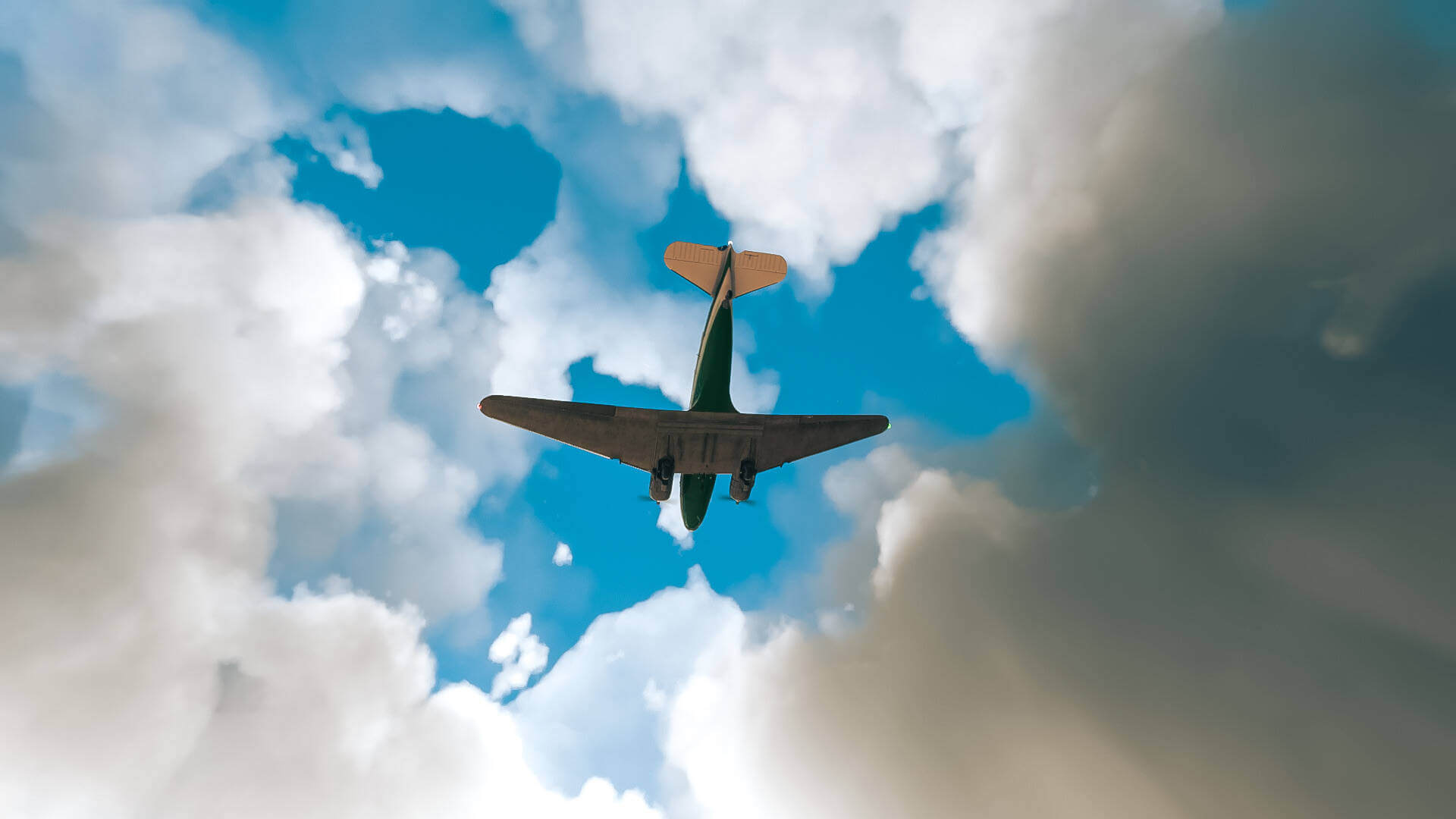 Bottom up view of a twin engine plane flying against a blue sky with white and gray cumulus clouds.