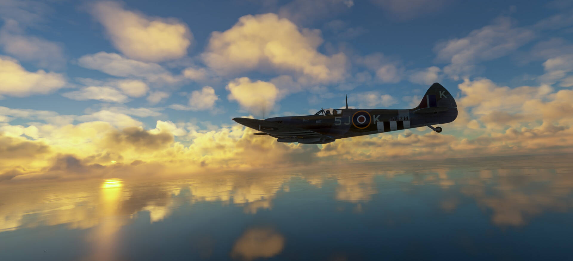 A Supermarine Spitfire flies low above a body of water. The clouds and sun are reflected in the water.