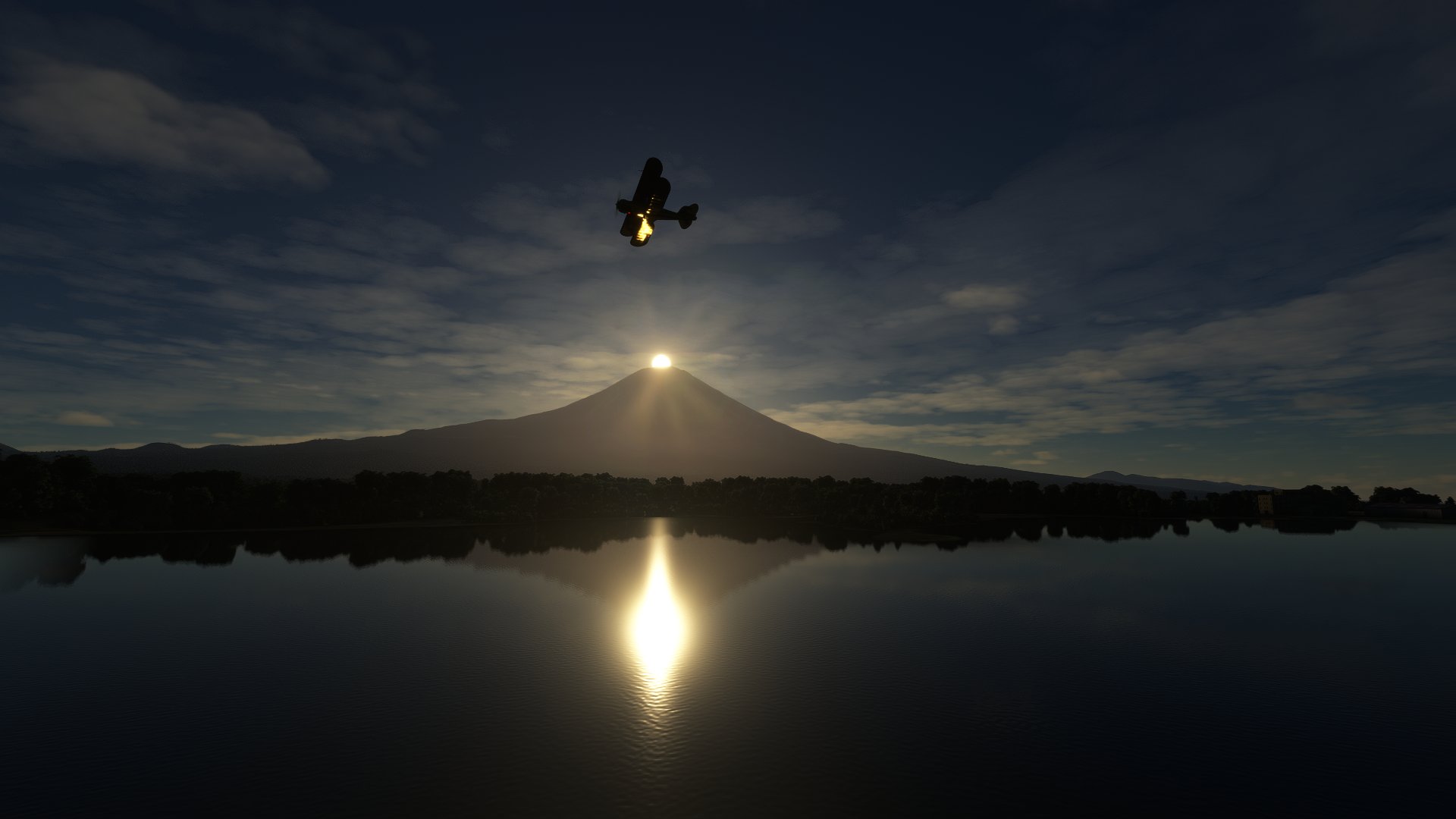 A Pitts Special flies over Mount Fuji in Japan. The sun is rising at the very top of the mountain. The sun is reflected in a lake below the plane.