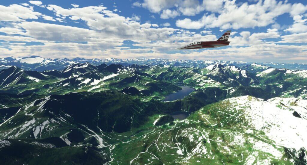 An L-39 Albatross flies over a green mountain valley. There is a lake in the distance.