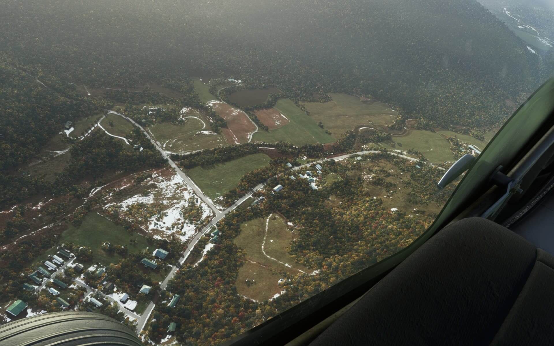 A pilot's perspective looking through the glass from their aircraft down below onto roads surrounded by fields, autumnal trees and scattered areas of snow and ice. 