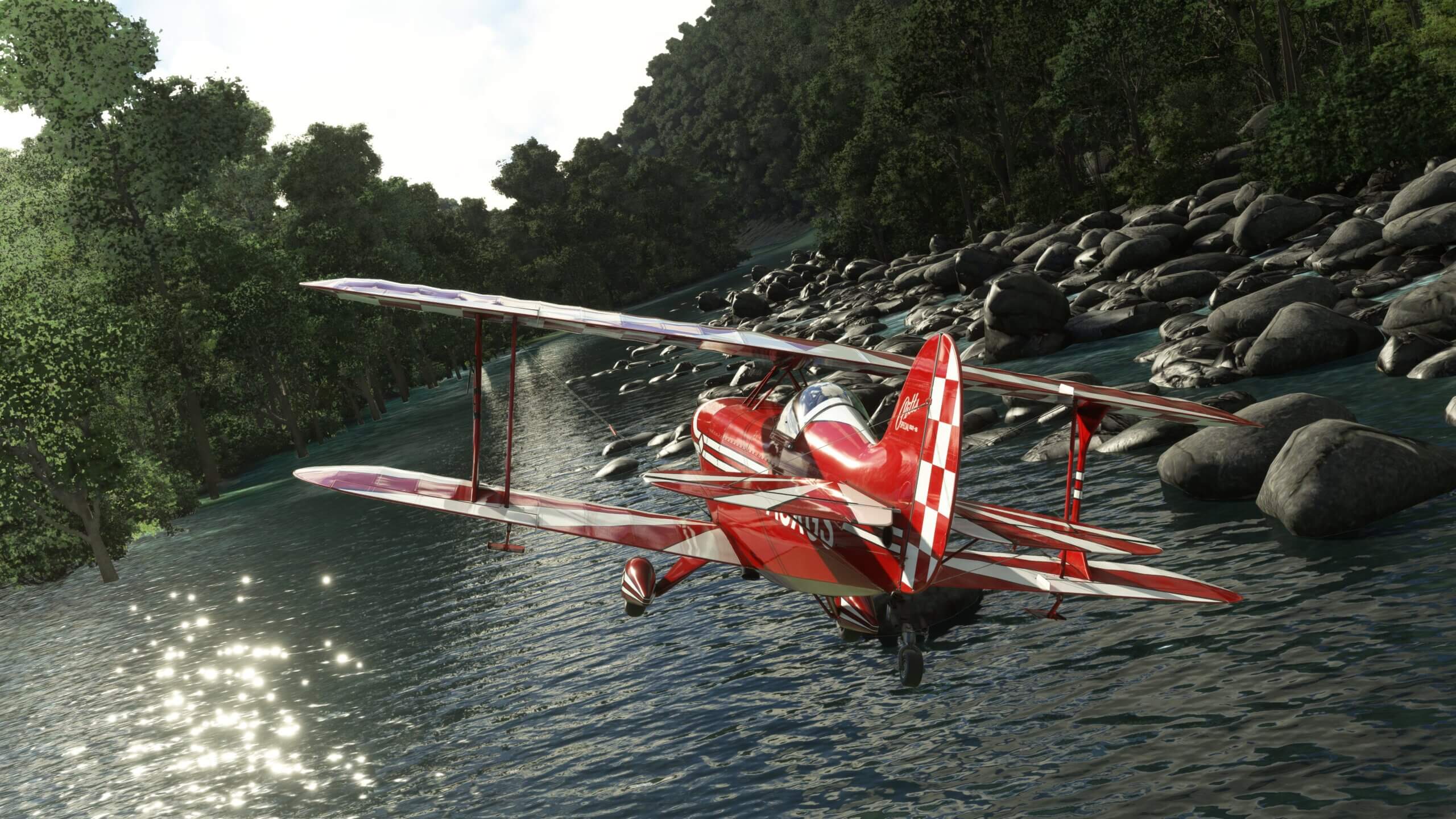A red and white bi-plane banks right over water with rocks close to the wingtips and trees either side of the water.