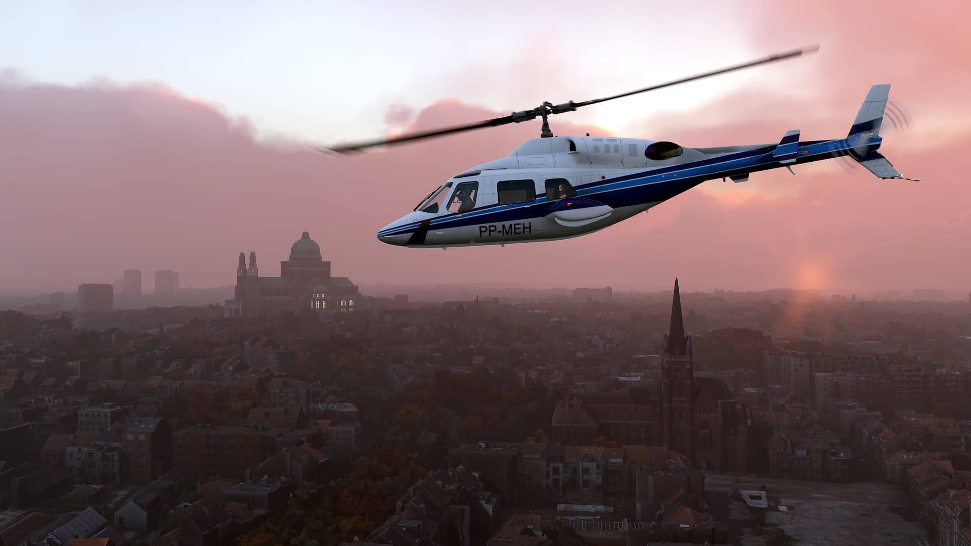 A Bell 407 helicopter passes over a city at dusk.
