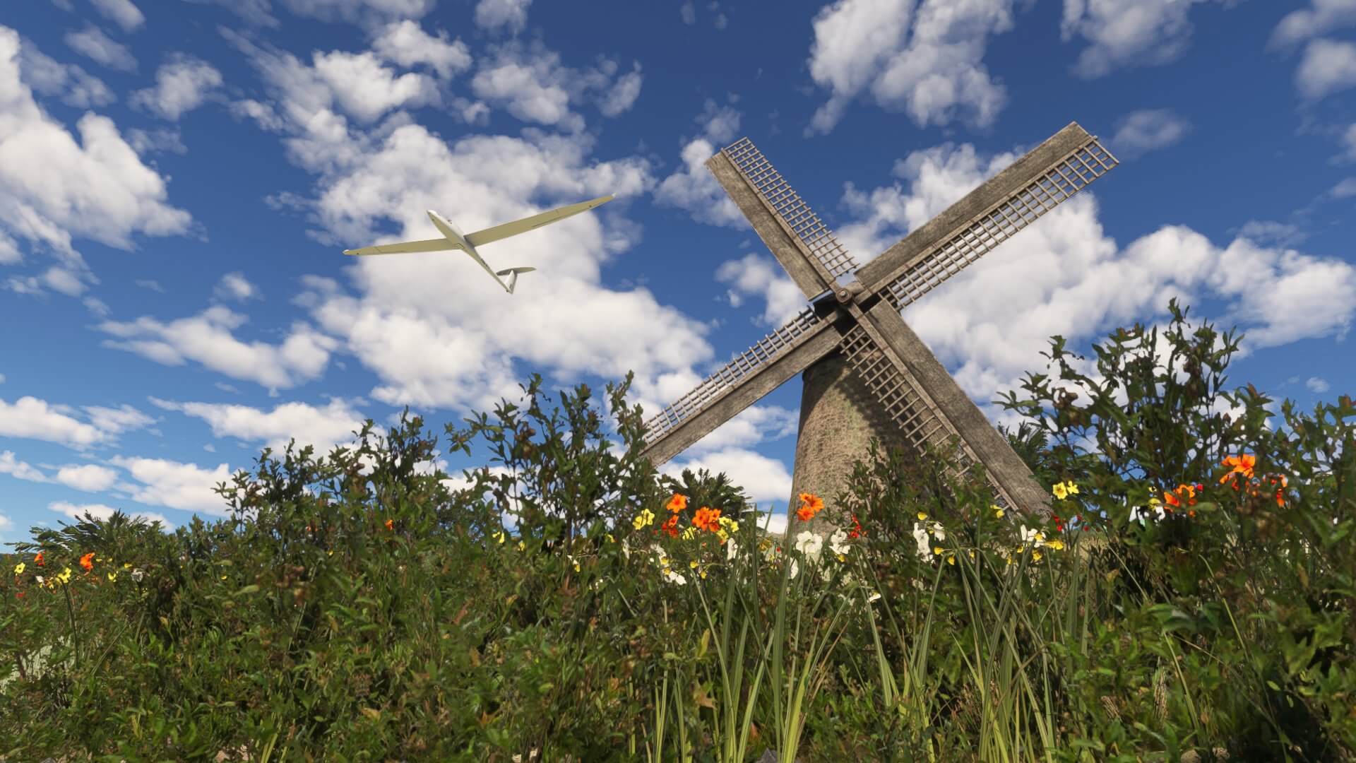 A glider passes above a windmill with flowers blooming in front.