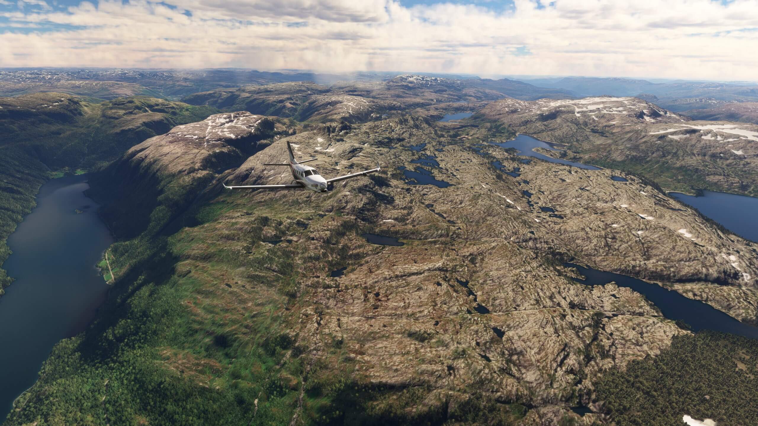The TBM 930 passes low over rocky terrain, with scattered clouds above