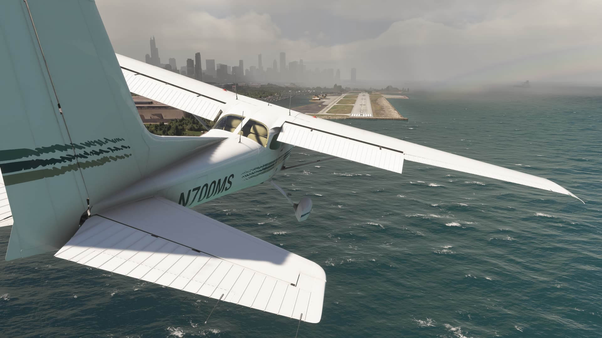 A Cessna 172 with flaps deployed banks to the right to correct its course whilst on short final to land on a runway strip ahead. The airport is surrounded by choppy water, with mist in the distance near city skyscrapers.