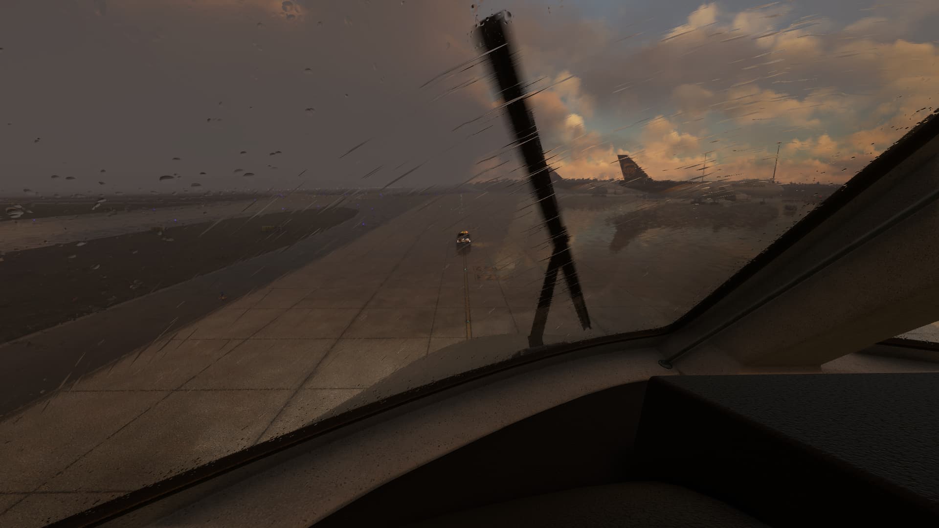 A forward facing view from the glareshield of an airliner, as the window wipers clear rain, and a ground crew vehicle on the apron ahead drives away.