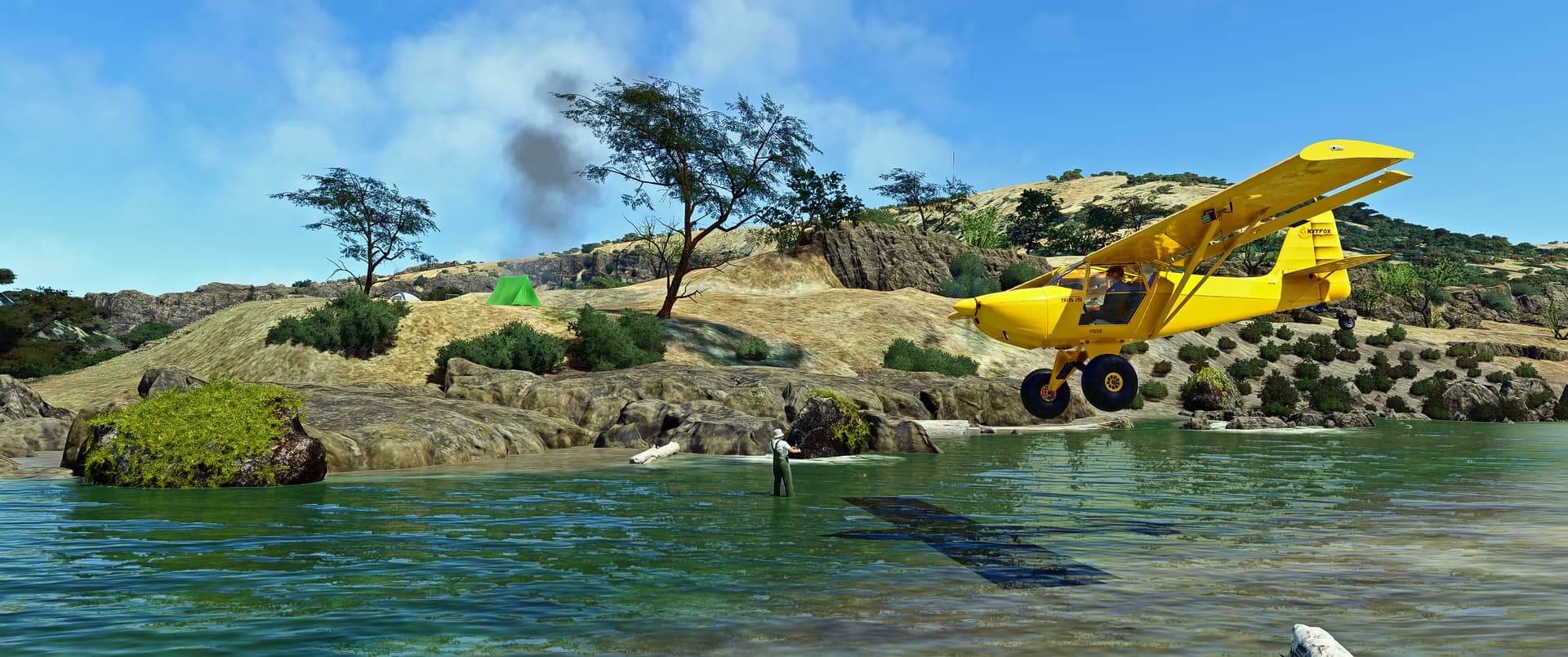 A yellow Kitfox passes very low over water next to a fisherman, who has a camping tent setup on the terrain above.