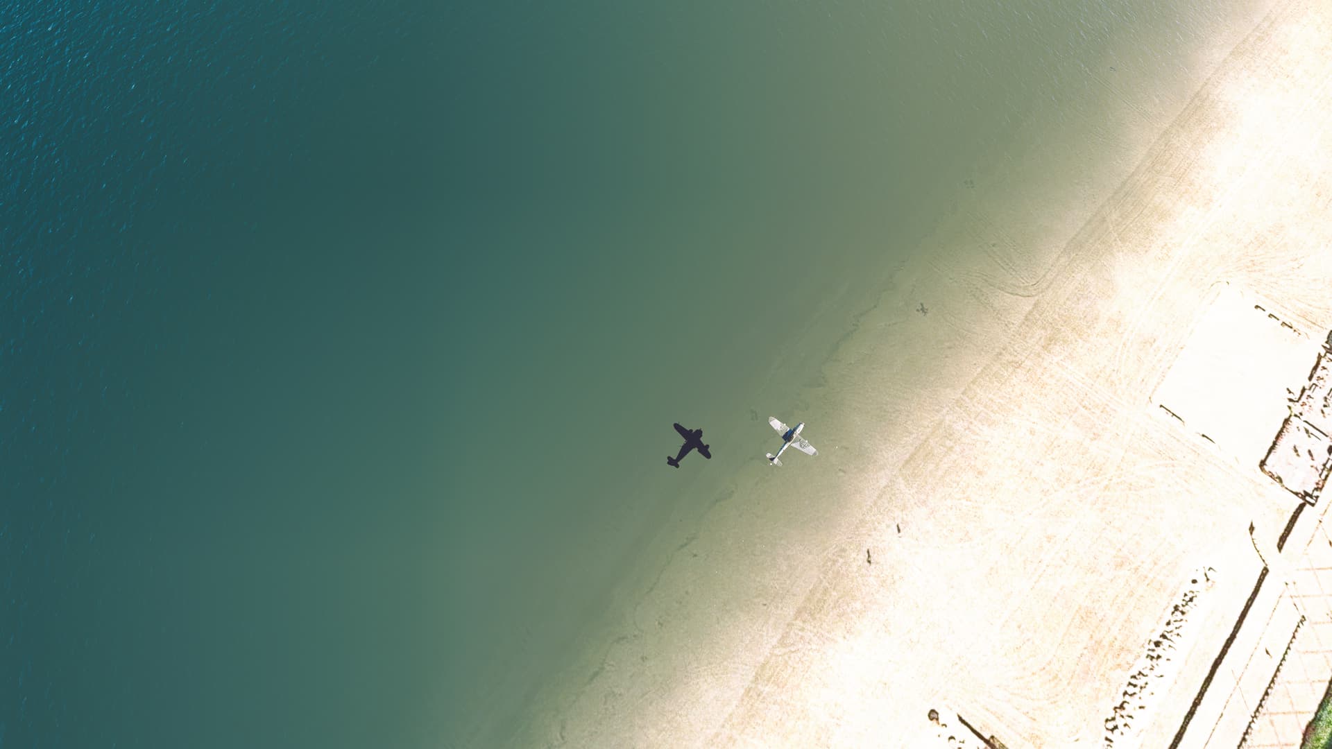 A low wing warbird aircraft casts a shadow on the sea below as it flies along a beach coastline from a birds eye view.