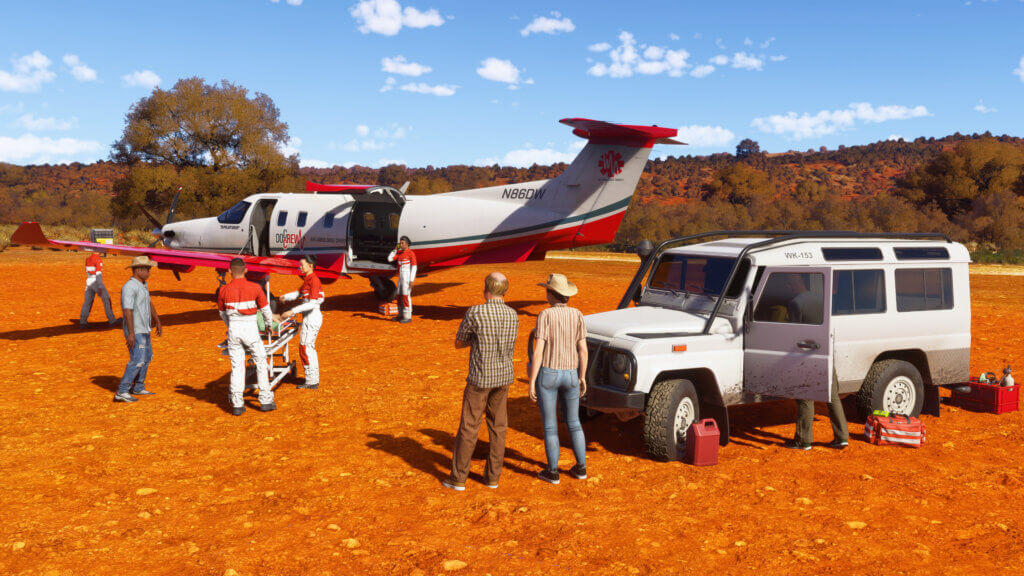 A red and white "DocCrew" Pilatus PC-12 is stationary on a desert strip whilst medical staff transfer a patient from a white 4x4 to the aircraft