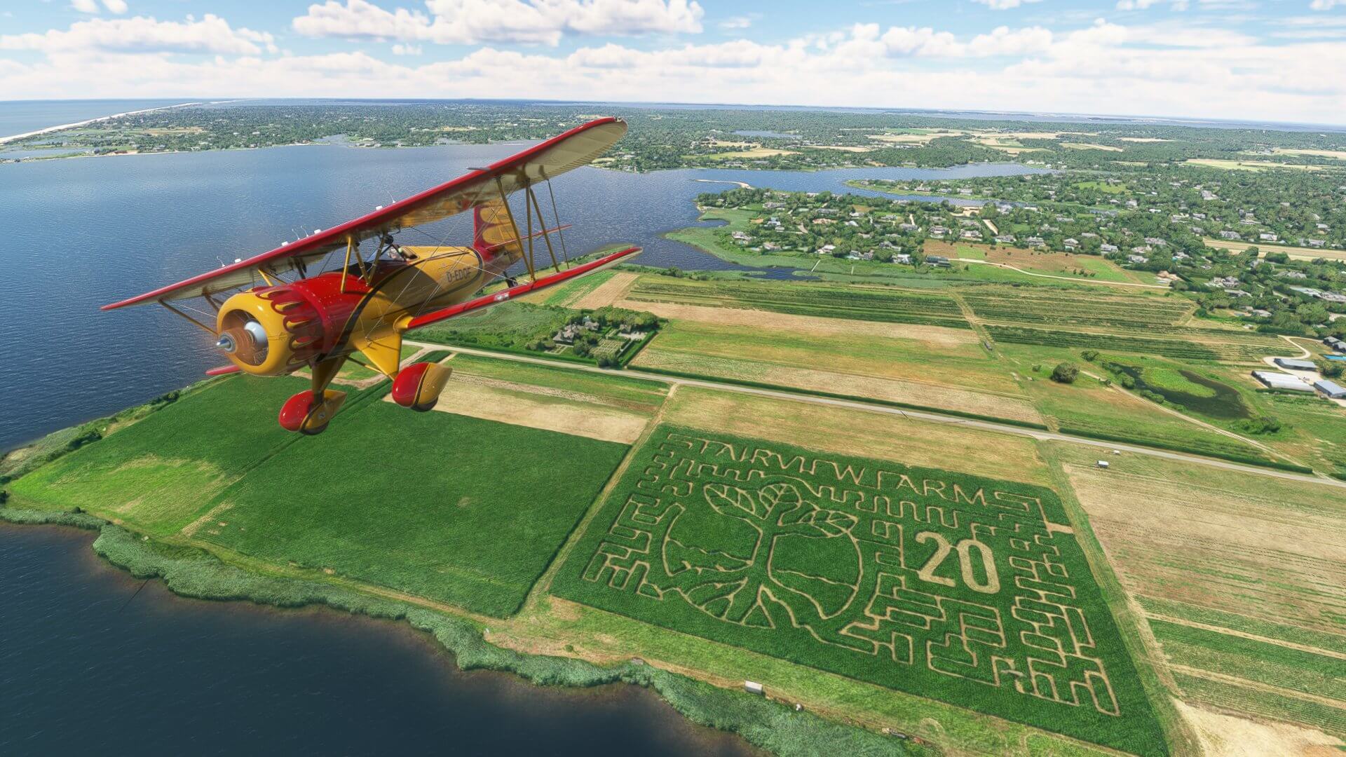 A dual-wing propeller aircraft in red and white paint flies above a field with patterns mowed into the crops.