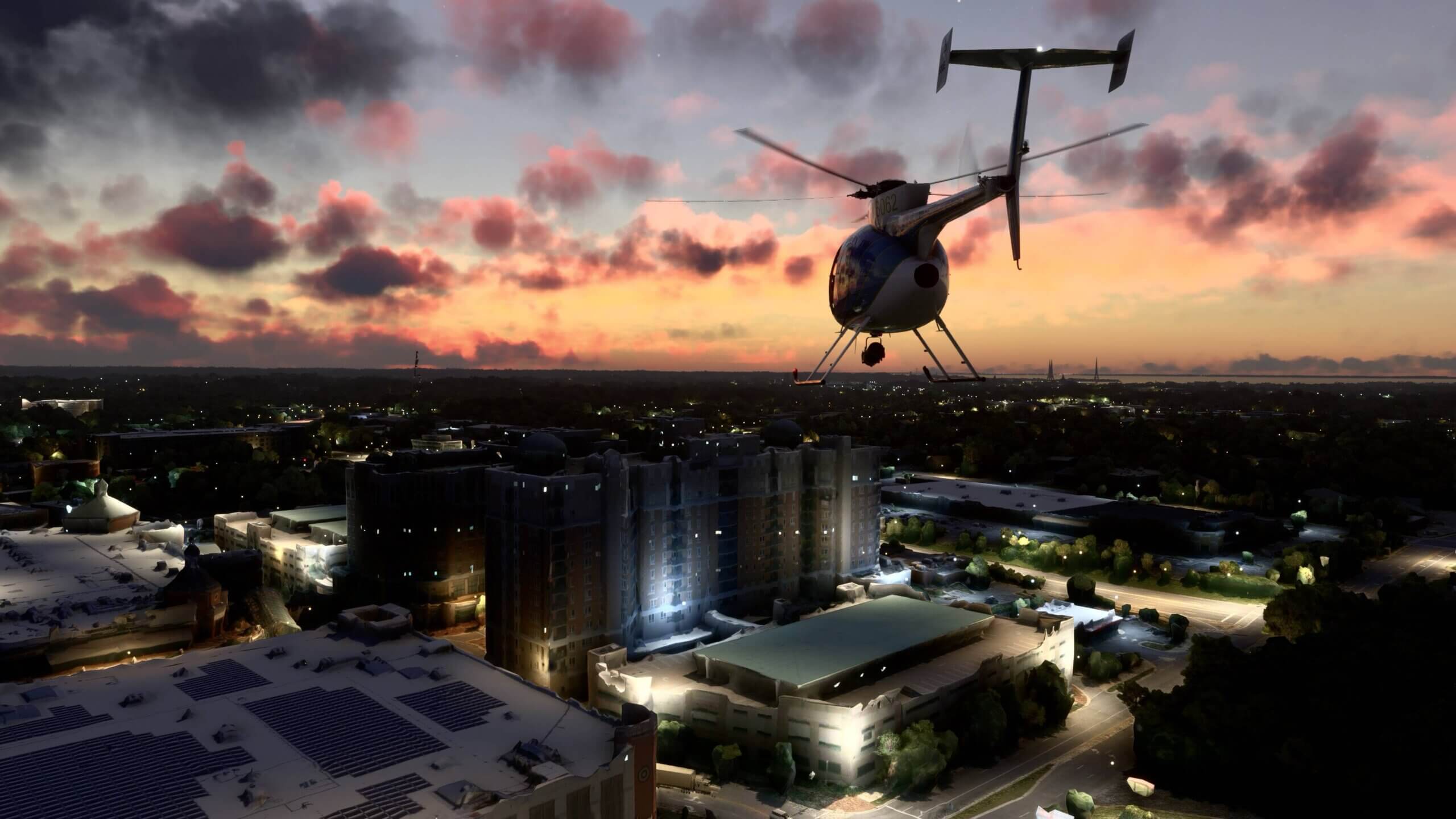 A small helicopter passes over a city with clouds above being lit pink from the setting sun