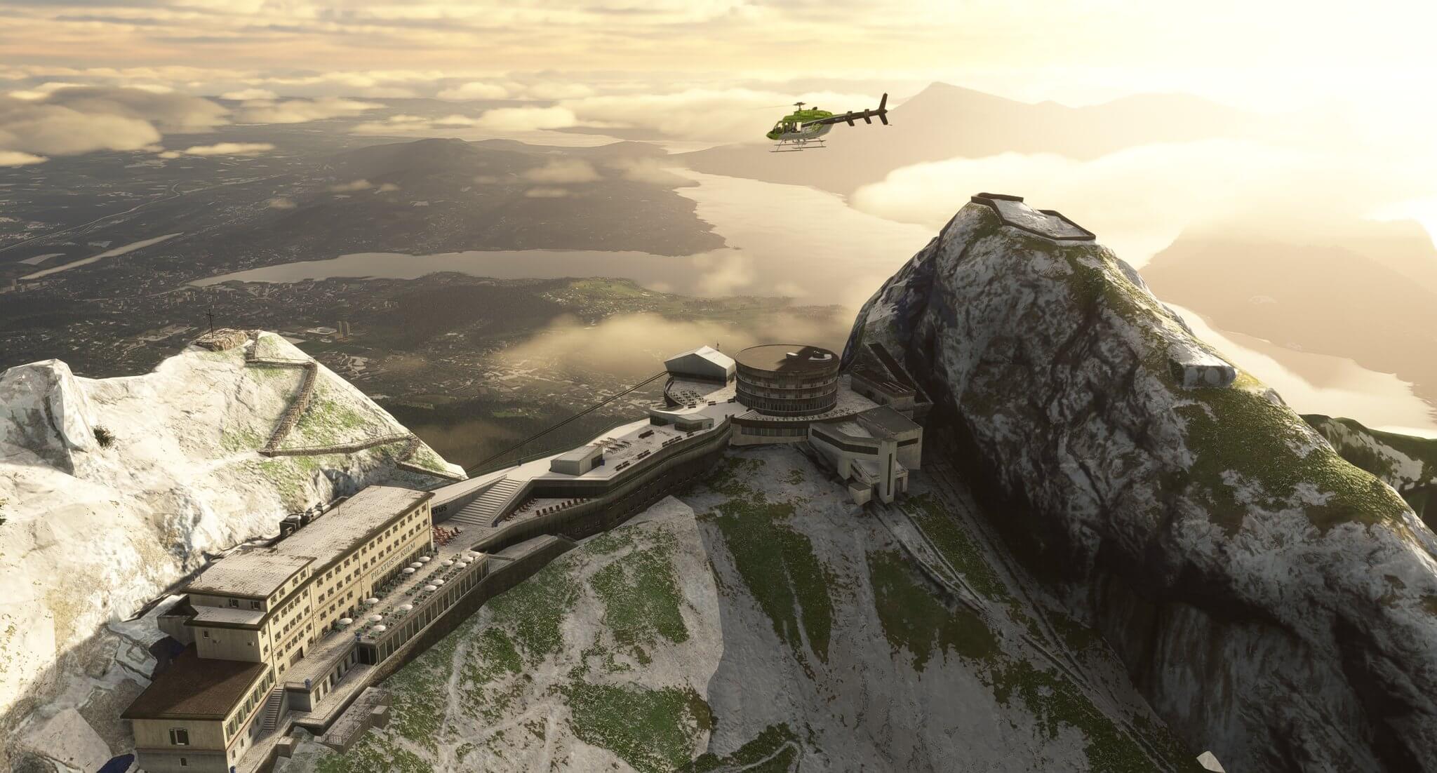 A Bell 407 flies over a set of buildings on top of a mountain range