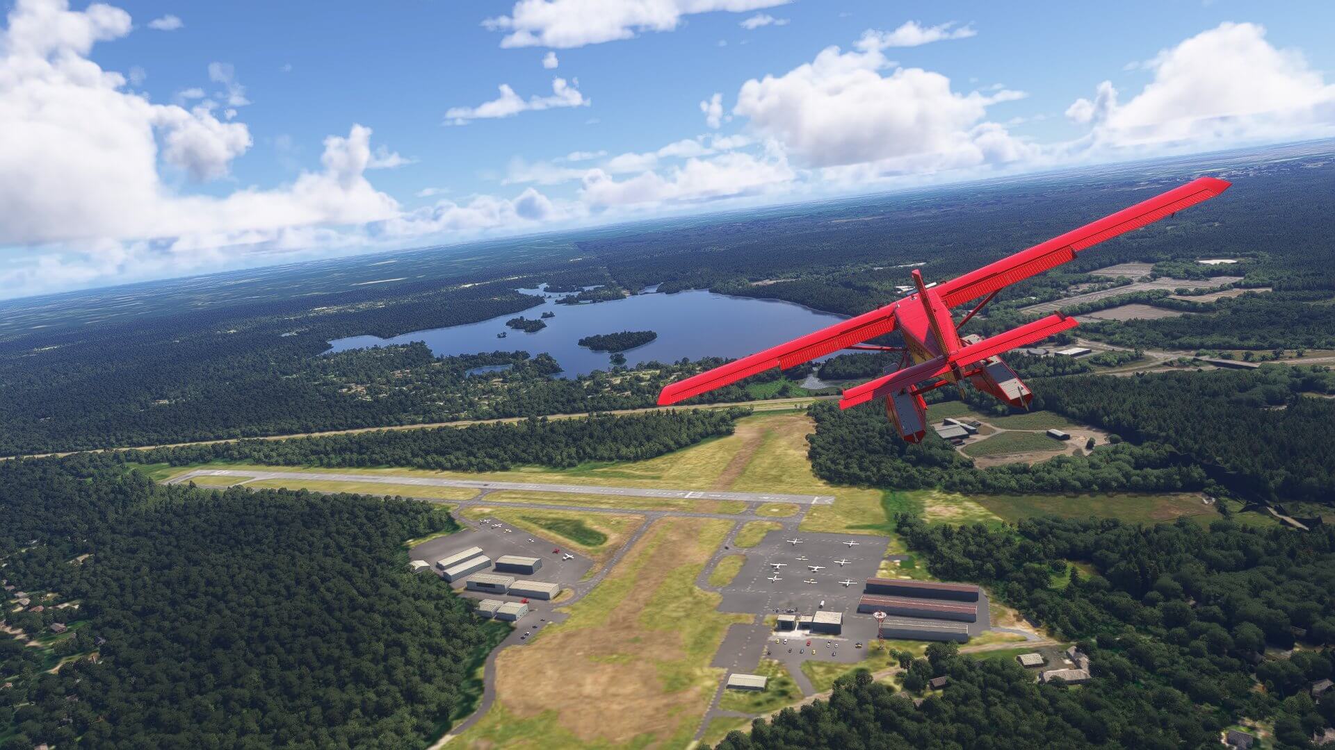A red high-wing propeller aircraft flies above an airfield with static planes in view on the apron below