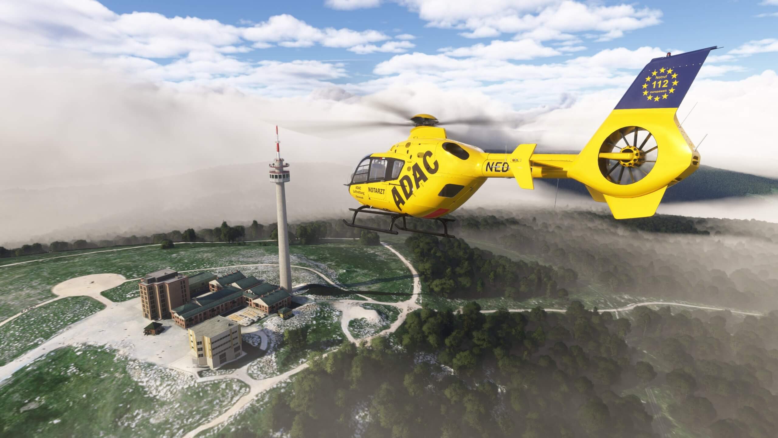 An ADAC EC-135 helicopter flies towards a tower with hazy clouds in the distance