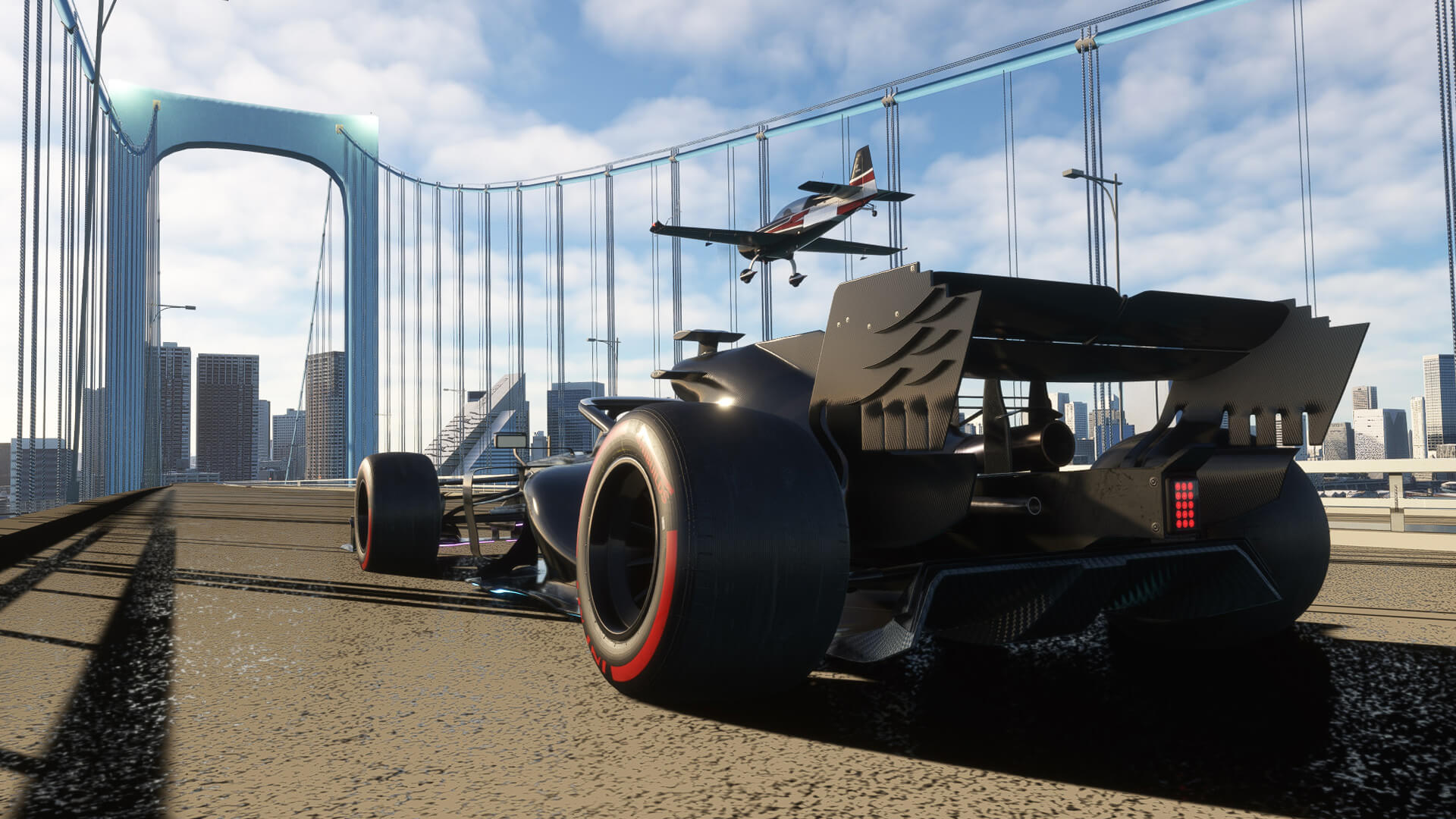An Extra 330 flies past a formula race car as they race across a bridge