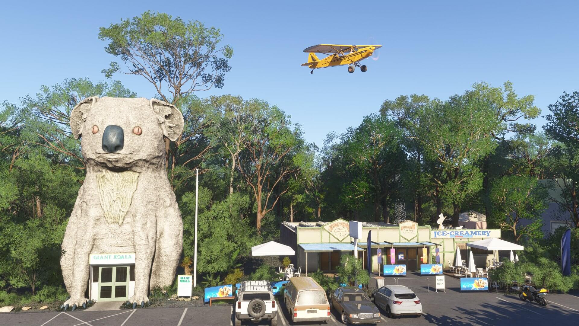 A high wing yellow prop aircraft flies over a giant Koala statue